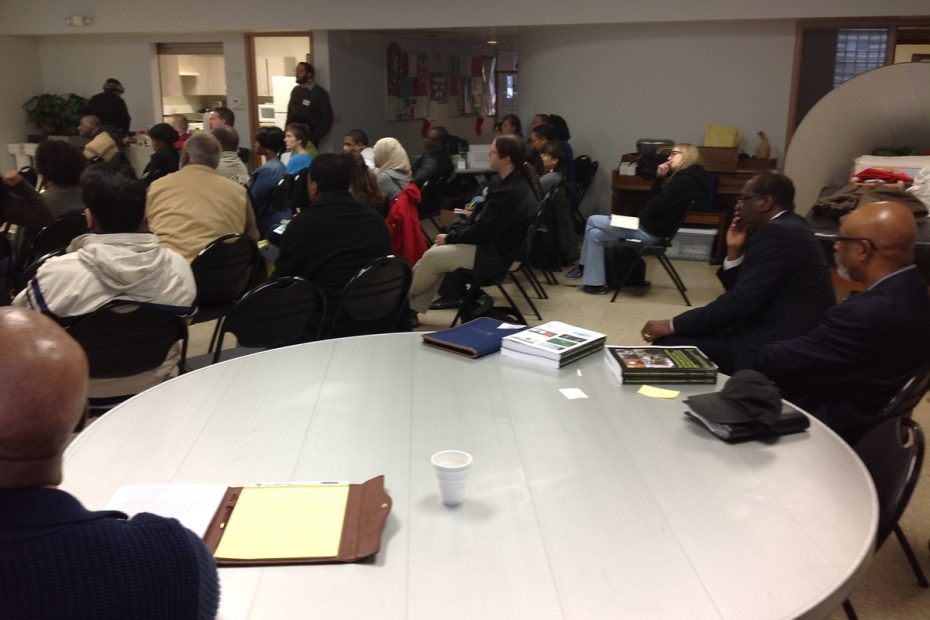 a community forum featuring a diverse crowd, some sitting in rows and other sitting at round tables.